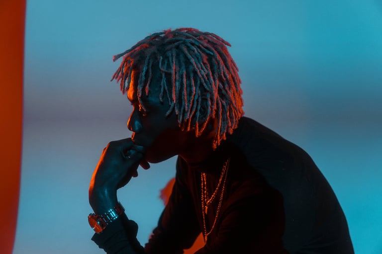 Person in profile wearing colorful dreadlocks and chains against a blue and red neon-lit background, hand near face in contemplative pose