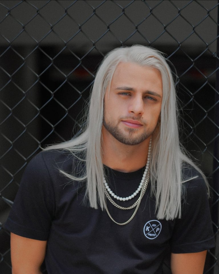 Man with long white hair and beard wearing black t-shirt with necklaces against chain-link fence backdrop